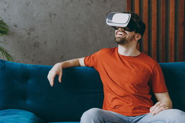 Wall Mural - Young happy man wears red t-shirt watch in vr headset pc gadget sit on blue sofa couch stay at home hotel flat rest relax spend free spare time in living room indoors grey wall. People lounge concept.