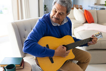 Happy Senior Biracial Man Playing Acoustic Guitar Sitting On Couch In Living Room At Home