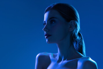 Portrait of female fashion model in neon light on blue gradient studio background. Beautiful caucasian girl with trendy make-up and well-kept skin.