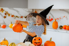 Girl In Witch Hat Posing With Coloured Pumpkins For Halloween On White Kitchen.