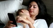 © Marco - Mother using cellphone device at home lying in bed with sleeping baby on chest napping