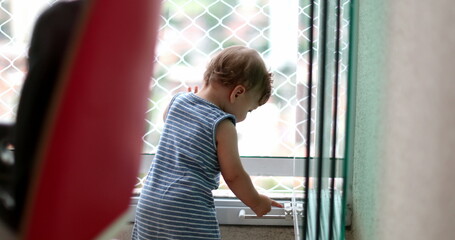 Cute baby standing by apartment window exploring home during lockdown, infant child staying at home