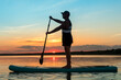A man in shorts and a T-shirt on a SUP board with a paddle against the backdrop of the sunset sky in the lake.