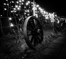 Old Carriage Wheel In Black And White Leaning Against A Wired Fence Decorated With Dangling Led Christmas Lights Looking Like Magic Stars Floating Just Above Ground.
