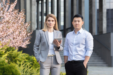 Portrait of two interracial businessmen. A young woman and a young Asian man are standing near the office in suit, Looking at the camera. A woman holds a tablet. A man holds his hands in his pockets.