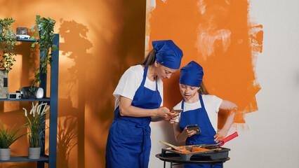 Canvas Print - Mother and girl looking at inspiration on mobile phone to paint house walls with orange color and paintbrush. Family painting interior using smartphone to find online decorating model. Tripod shot.