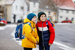 Two little kids boys of elementary class walking to school during snowfall. Happy children having fun and playing with first snow. Siblings and best friends with backpack in colorful winter clothes.