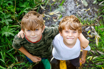 Wall Mural - Two happy little school kids boys, funny siblings having fun together walking through water in river in gum rubber boots. Family portrait of healthy brothers and best friends