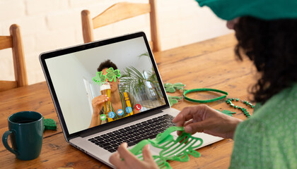 Sticker - Mixed race woman making st patrick's day video call to friend drinking beer on laptop at home