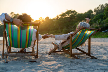 Happy Asian family senior couple enjoy outdoor lifestyle on summer beach holiday vacation at the sea. Elderly retired man and woman relaxing on beach chair and holding hands together at summer sunset