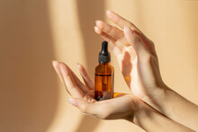 Women's Hands Hold Bottle Of Amber Glass With Cosmetic Serum On Brown Background. Mockup Of Container With Dropper Lid With Collagen In Rays Of Sunlight. Concept Of Skin Care