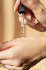 Wall Mural - Close-up of pipette with collagen moisturizing hyaluronic serum in woman's hand in rays of sunlight. Vertical image beige background