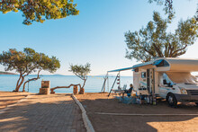 Family Traveling With Motorhome Are Eating Breakfast On A Beach. Travelers On An Active Family Vacation With Motorhome RV Parked On The Beach Under A Tree Facing The Sea, Crete, Greece.