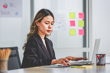 Millennial Asian cheerful successful professional female businesswoman sitting smiling using tablet notebook computer at working with document folder in company office workstation.