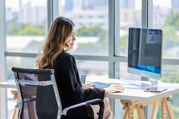 Millennial Asian cheerful successful professional female businesswoman sitting smiling using typing computer keyboard at working desk near windows with document folder in company office workstation