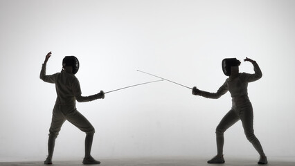 Side view of two young female swordswoman with crossed rapiers. Athletes in uniforms and protective helmet mask fighting a duel in dark studio on white background. Girls holding epees and attack.