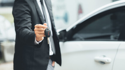 A man in a suit stands holding the car keys next to a white car, getting a new car with a car showroom dealer. Car trading concept.