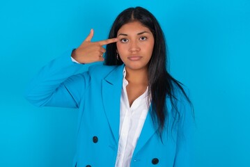 Wall Mural - Unhappy Young latin woman wearing  blue blazer blue background makes suicide gesture and imitates gun with hand, curves lips keeps two fingers on temple, shoots, being tired of everything,