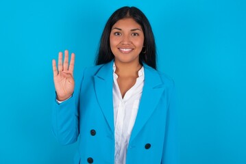 Wall Mural - Young latin woman wearing  blue blazer blue background showing and pointing up with fingers number four while smiling confident and happy.