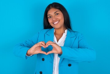 Wall Mural - Young latin woman wearing  blue blazer blue background smiling in love doing heart symbol shape with hands. Romantic concept.