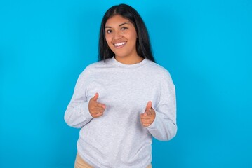 Young latin woman wearing gray sweater blue background directs fingers at camera selects someone. I recommend you. Best choice