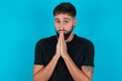 © Jihan - Positive young bearded hispanic man wearing black T-shirt over blue background smiles happily, glad to receive pleasant news from interlocutor, keeps palms together, People emotions concept.