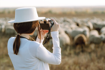 Young woman with a camera in front of a herd of sheep.  Fashion, style concept. People, lifestyle, relaxation and vacations concept.