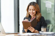 © Phushutter - Happy asian business woman holding notebook paper and working with laptop in formal wear sitting at office desk. reading report document.
