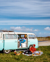 Old Vintage Camper Van Parked By A Lake With A Couple Of Men And Women Watching The Sunrise Over The Meadow During Spring Season, Couple Picnic In The Park During Spring, Camper Van