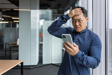 Asian businessman inside office building upset and sad reading bad news on mobile phone, business owner investor using smartphone standing near window