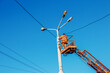 A municipal worker in protective equipment performs hazardous work to eliminate an interruption in the power grid. A worker repairs a street lamp from an aerial platform basket.