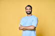 Smiling confident bearded indian man standing isolated on yellow background. Happy ethnic guy wearing blue t-shirt looking at camera posing with arms crossed for portrait.