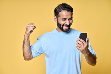 Excited overjoyed happy indian man raising fist screaming using smartphone winning mobile game, betting money prize holding cell phone celebrating victory isolated on yellow background.