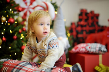 Happy little toddler boy playing with Chistmas lights in a cozy living room on Christmas eve. Celebrating Xmas at home.