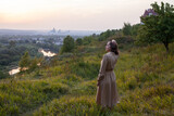 Fototapeta Konie - Beautiful young smiling girl in a long brown dress stands along the lawn. Happy woman walks at sunset on a hill overlooking the river. Concept of having rest in park during summer holidays or weekends