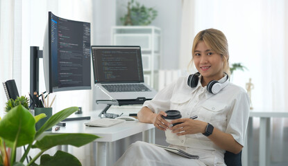 Female programmers sitting front of laptop and large curved computer monitor with programming code on screen and smiling to camera.