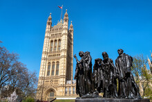 The Burghers Of Calais Statue, By Auguste Rodin, Completed In 1889, Outside Of The Houses Of Parliament, London, UK. The Victoria Tower Has A Union Jack Flag Flying Against A Blue Sky