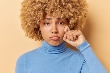 Wall Mural - Photo of dejected frustrated woman wipes tears feels very upset after hearing bad news watches melodrama recalls unhappy memory wears blue poloneck poses against beige background. Negative emotions