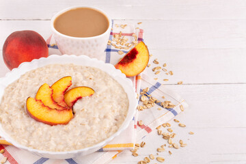 Wall Mural - Oatmeal porridge with peach in a bowl (plate) and coffee in a cup on a white wooden background. Place for text, recipe. Healthy breakfast.