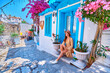 Girl traveler wearing dress and hat walks on beautiful colorful flower street with white houses and blue doors in a European city