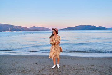 Wall Mural - Serene calm young pensive girl traveler wearing hat and dress stands alone and looking into the distance at summertime