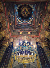 Interior Architectural Details Of The Curtea De Arges Monastery. The Hall With Tall Columns, Christian Orthodox Painted Icons On Walls And A Golden Chandelier With Lights Suspend Out Of Ceiling