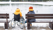 Caucasian Woman In A Wheelchair And Her Friend Are Sitting By The Lake With Ducks In Winter.