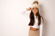 © Danko - Pretty brunette kid girl wearing white winter hat over white background Doing peace symbol with fingers over face, smiling cheerful showing victory
