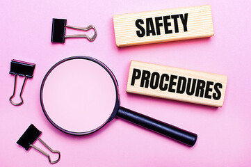 On a pink background, a magnifier, black paper clips and wooden blocks with the text SAFETY PROCEDURES. Business concept