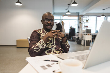 A black African American businessman in a national ethnic business suit works at a laptop. Analytics and work online