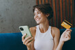 © ViDi Studio - Young minded woman wear white tank shirt using mobile cell phone credit bank card shopping online booking tour sit on blue sofa stay home flat rest relax spend free spare time in living room indoors.
