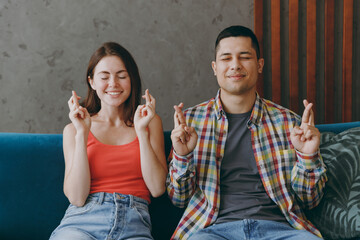 Young couple two friends family man woman in casual clothes wait special moment keep fingers crossed sits on blue sofa together stay at home rest spend free spare time in living room indoor grey wall