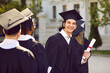 Happy young man having fun at his graduation. Outdoor portrait of a cheerful, joyful university or college graduate in his cap and gown standing among other students, holding his diploma and smiling