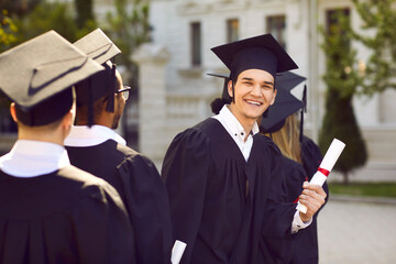 Wall Mural - Happy young man having fun at his graduation. Outdoor portrait of a cheerful, joyful university or college graduate in his cap and gown standing among other students, holding his diploma and smiling